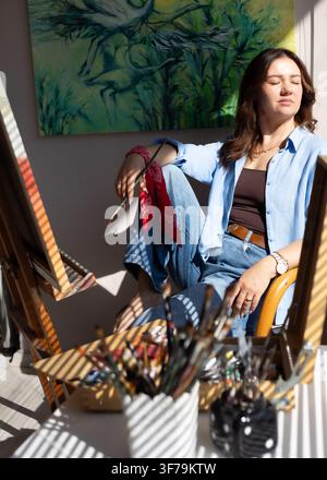 Portrait d'une artiste féminine se détendant à côté de son chevalet dans un atelier lumineux rempli d'une lumière naturelle chaude. Banque D'Images
