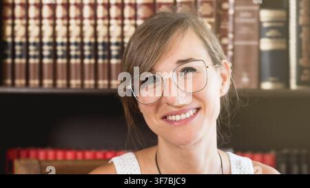 Portrait d'une jeune femme souriante avec des lunettes à la maison devant une étagère à livres. Image lifestyle chaleureuse pour la confiance, le bien-être, le bureau à domicile et l'amitié Banque D'Images