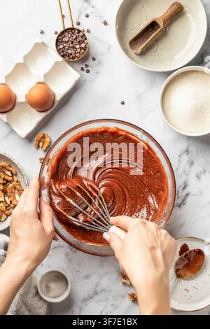 Procédé de fabrication du brownie au chocolat. Mains féminines fouettant la pâte dans un bol en verre, vue de dessus. Concept de pâtisserie maison, recette Banque D'Images