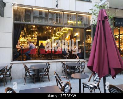 Une scène de terrasse de restaurant en plein air au crépuscule, avec un bar ouvert sur le devant doté de grandes fenêtres et portes en verre. À l'intérieur, les clients sont assis à des tables avec du rouge Banque D'Images