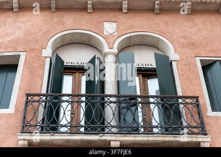 ITALIE, VENISE, 11 juillet 2025 : Une façade en stuc pastel avec des fenêtres en arc, des volets verts, une rambarde de balcon en fer et une enseigne Prada visible est montrée depuis Banque D'Images