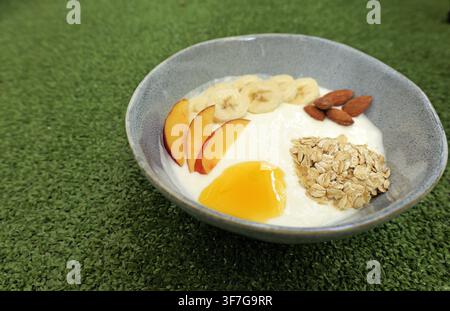 Bol de petit-déjeuner sain mettant en vedette un yaourt crémeux garni de bananes tranchées, de tranches de pêches fraîches, d'avoine roulée, de miel doux et d'amandes entières, créant un Banque D'Images
