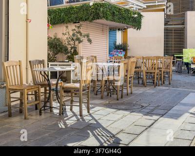 Des chaises en bois et de petites tables remplissent un café de trottoir vide sous une verdure feuillue sur une allée piétonne ensoleillée à Monastiraki, Athènes Banque D'Images