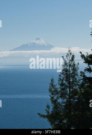Volcan Osorno distant, coiffé de neige, émergeant au-dessus des nuages bas sur le lac Llanquihue, encadré par des arbres, Los Lagos, Chili. Banque D'Images