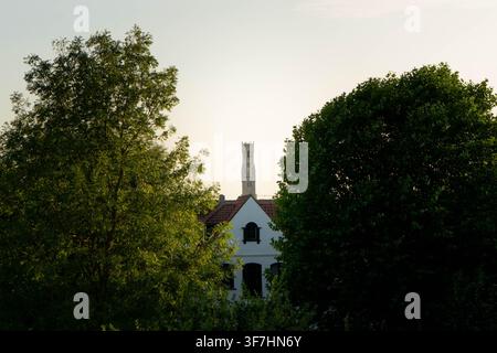 BRUGES, BELGIQUE – 9 AOÛT 2026 : Le beffroi médiéval de Bruges visible en arrière-plan encadré par des arbres verts et une maison blanche traditionnelle Banque D'Images