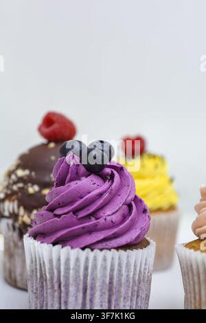 Un cupcake violet avec un glaçage en spirale et des myrtilles se tient, est exposé et présenté parmi des desserts colorés, des douceurs sucrées, de la nourriture de fête. Banque D'Images