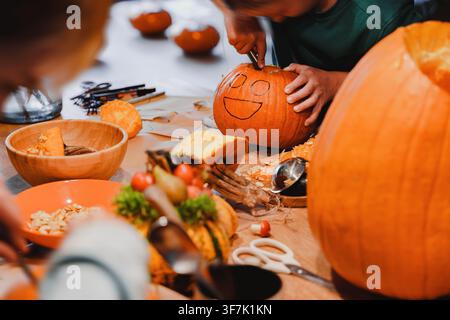 Enfant découpe soigneusement un visage souriant dessiné sur un citrouille orange lors d'une activité créative de sculpture de citrouille d'Halloween, citrouille d'Halloween ca Banque D'Images