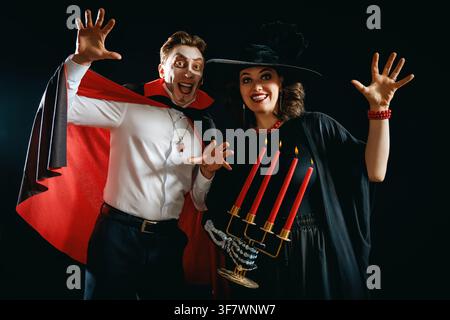 Couple adulte joyeux en costumes d'Halloween dramatiques posant avec enthousiasme devant un fond sombre. L'homme porte une tenue classique de vampire avec un re Banque D'Images