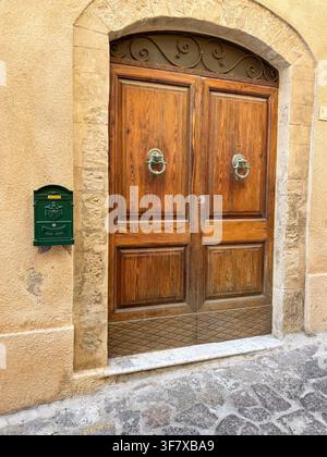 Porte en bois rustique avec poignée en métal ornée, flanquée par une arche en pierre sur une rue pavée. Une boîte aux lettres verte est fixée à côté, rehaussant la Banque D'Images