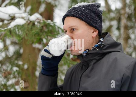 Homme barbu buvant du thé chaud dans une tasse de voyage en métal dans une forêt enneigée en hiver. Homme mûr à la barbe courte savourant une boisson chaude lors d'une paisible promenade en plein air parmi des arbres à feuilles persistantes couverts de neige Banque D'Images