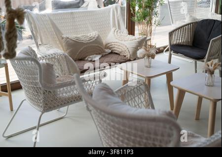 Espace de détente en terrasse baigné de soleil avec chaises tressées, canapé assorti, tables basses en bois, sol carrelé clair, coussins neutres, couverture tricotée, verdure douce Banque D'Images