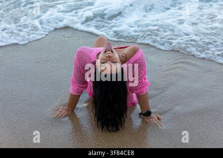 Femme souriante en pull rose profite de la plage, adossée sur le sable mouillé avec des vagues de l'océan en arrière-plan. Banque D'Images