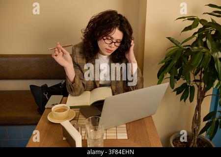 Freelance aux cheveux bouclés s'engage dans des projets en ligne à une table confortable de café Banque D'Images