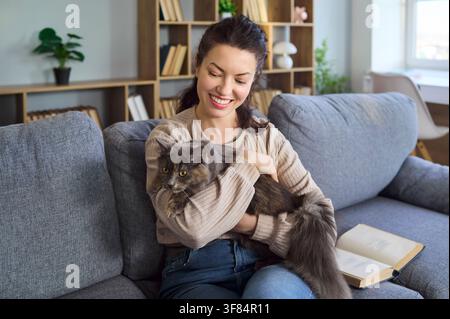 Femme souriante câlinant son chat sur le canapé à la maison, style de vie confortable de propriétaire d'animal de compagnie Banque D'Images