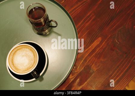 Cappuccino frais et un verre de thé servis ensemble sur une table ronde de café. Boissons chaudes dans un cadre cosy de café. Banque D'Images