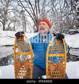 Homme âgé avec des raquettes en bois classiques (Bear Paw) sur une rive de ruisseau, paysage d'hiver au Colorado Banque D'Images