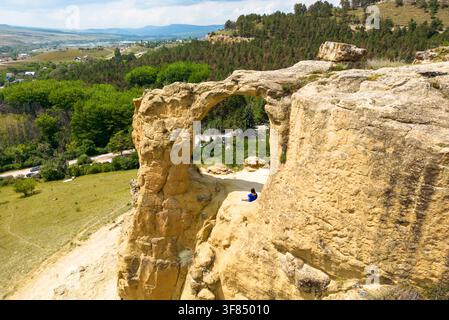 Une vue aérienne d'une grande formation d'arche de pierre naturelle sur une colline. Une femme portant une robe bleue est assise sur la corniche rocheuse sous l'arche, regarde Banque D'Images