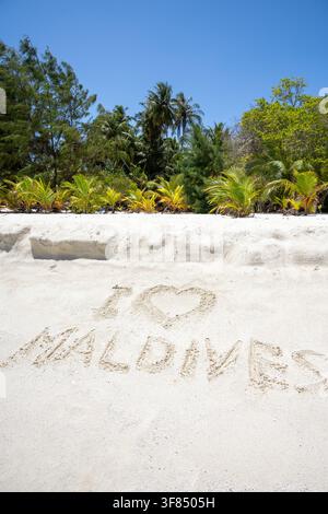 Une photographie verticale d'une plage de sable blanc avec la phrase Je t'aime les Maldives dessinée dans le sable au premier plan, utilisant un symbole de cœur pour le mot l Banque D'Images