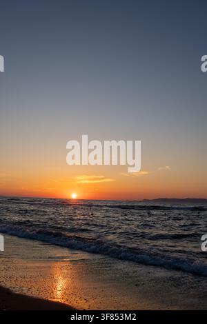 Le soleil touche l'horizon de la mer en projetant une lumière orange sur les vagues ondulantes. Vue côtière avec rive de plage mouillée, réflexion du coucher de soleil sur le sable, ciel dégradé et distan Banque D'Images