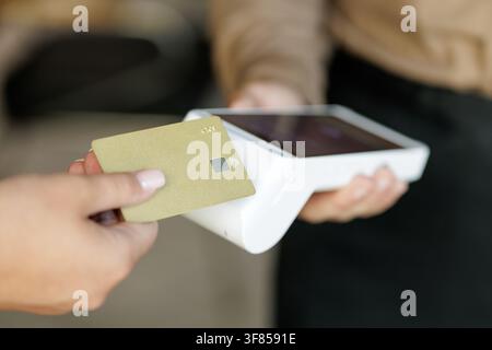 Gros plan d'un client tapant une carte bancaire sans contact sur un terminal POS sans fil moderne, illustrant le paiement par carte NFC, le paiement sans espèces et la fintech en Banque D'Images