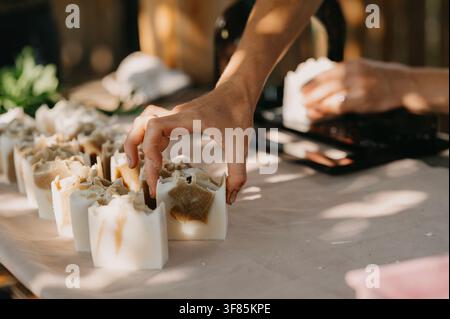 Main posant une savon fait main sur une table de marché rustique sous une lumière chaude du soleil avec des ombres douces, texture marbrée crémeuse Banque D'Images