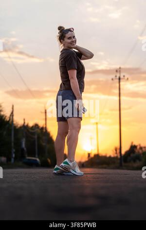 Femme debout sur une route souriant et posant avec la main dans les cheveux, dans une scène de coucher de soleil avec des lignes électriques et des arbres, avec route, lignes électriques, arbres, ciel Banque D'Images