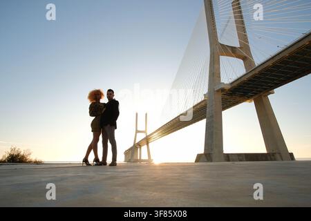 Couple posant sur un sol en béton avec le pont Vasco de Gama en arrière-plan pendant le coucher de soleil Banque D'Images