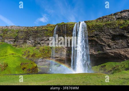 Vue panoramique du célèbre cascade de Seljalandsfoss en Islande par une journée ensoleillée, mettant en scène un arc-en-ciel éclatant dans la brume d'eau. Banque D'Images