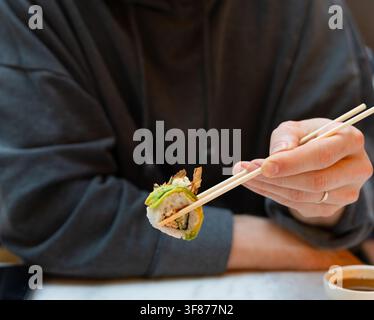 Un gros plan de mains prenant des sushis avec des baguettes dans un moment de concentration au restaurant. La technique précise démontre une habileté exercée. Chaque mouvement reflète le soin apprécié dans la culture alimentaire japonaise Banque D'Images
