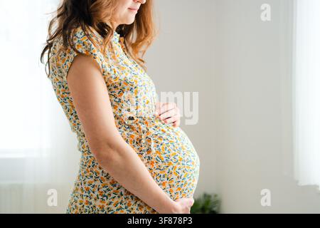Une femme enceinte en robe fleurie se tient de profil en embrassant son ventre dans un studio. L'élégante mère caressant son ventre montre une protection tendre et aimante ainsi qu'un calme serein et paisible à l'intérieur. Banque D'Images