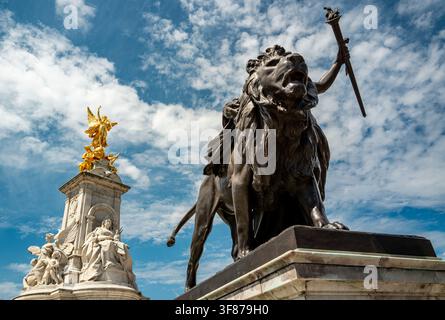 Sculpture d'un lion en bronze au Victoria Memorial à Londres, avec la reine Victoria et une Victoire ailée dorée sous un ciel nuageux. Banque D'Images