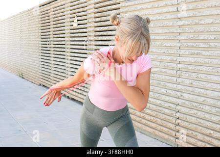 Femme âgée effectuant un swing de kettlebell en plein air sur un passage piétonnier urbain avec une expression concentrée, une posture forte, un haut rose, un legging gris et une clôture ensoleillée Banque D'Images