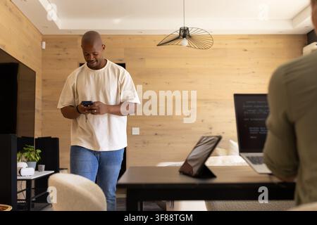 Couple divers habillé de manière décontractée, debout et travaillant à la maison en utilisant un smartphone, un ordinateur portable et une tablette. Espace de travail, moderne, minimaliste, salon, duo, rem Banque D'Images