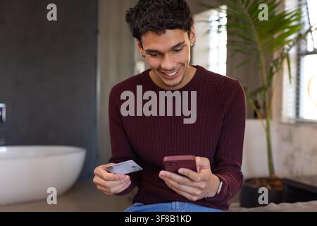 Homme afro-américain assis sur le rebord de la baignoire, en pull marron, souriant, vérifiant son téléphone avec carte. Salle de bain, moderne, indépendante, plante, smartphone, sur Banque D'Images