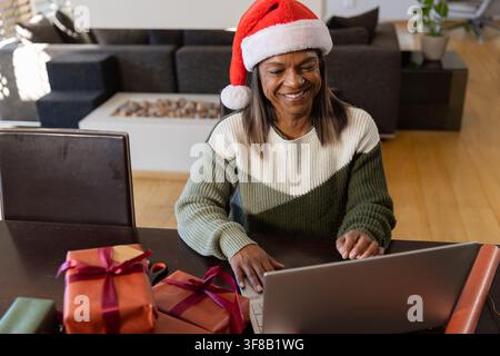 Femme afro-américaine mûre avec chapeau du Père Noël et pull en tricot emballant des cadeaux à la maison en utilisant un ordinateur portable. Festif, confortable, vacances, télétravail, lumière du jour, vert Banque D'Images