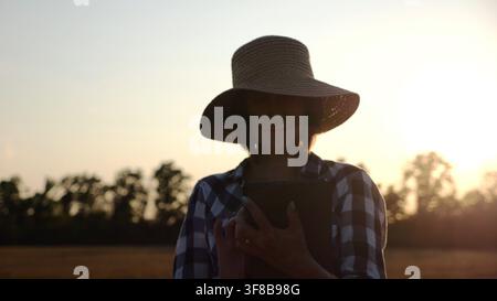 Agronome femelle en chapeau de paille surveillant la récolte sur le champ d'orge au coucher du soleil. Agriculteur adulte utilisant la tablette numérique sur la prairie de blé au crépuscule. Magnifique Banque D'Images