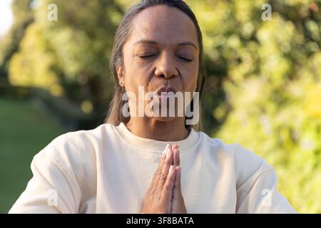 Femme afro-américaine debout dans un jardin baigné de soleil, joignant les paumes en prière, portant un sweat-shirt crème. Méditation, pleine conscience, verdure, espace vert, ou Banque D'Images