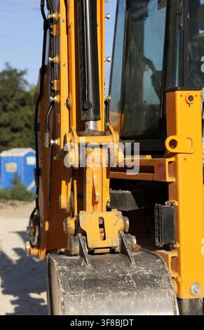 Détail du bras d'excaveur jaune montrant le cylindre hydraulique et la benne sale, inactif sur un chantier poussiéreux avec une cabine de toilette portable dans le Banque D'Images