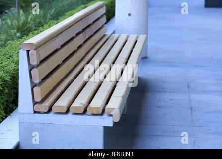 Banc à lattes en bois reposant sur une base en béton, offrant des sièges dans un parc urbain contemporain ou une zone de jardin avec un feuillage vert et un gr pavé Banque D'Images