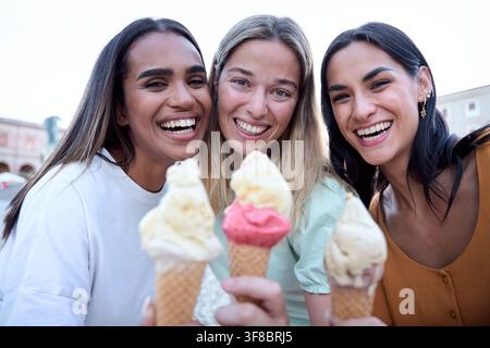 Trois amies heureuses prenant un selfie tout en mangeant de la crème glacée sur une place de la ville Banque D'Images