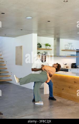 Couple afro-américain dansant et plongeant dans une cuisine ouverte près d'un îlot en bois avec une lumière chaude. Minimaliste, moderne, contemporain, spacieux, bois Banque D'Images