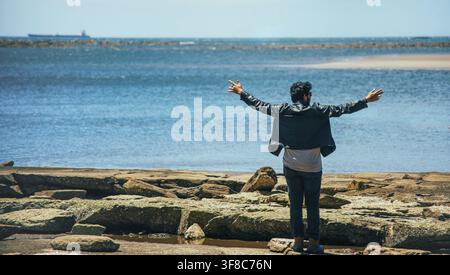 Homme debout sur le rivage rocheux, les bras écartés, regardant l'océan. Homme dos à l'appareil, les bras écartés, regardant au loin Banque D'Images
