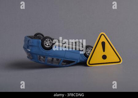 Voiture-jouet renversée à côté d'un panneau d'avertissement sur fond gris Banque D'Images