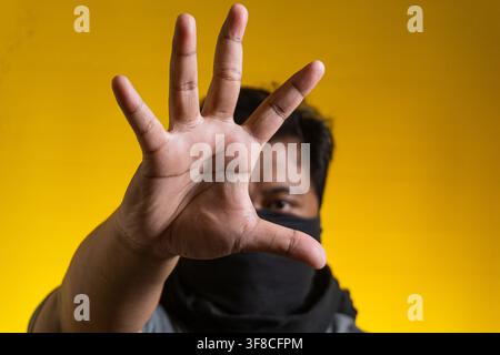 Portrait frontal d'un jeune homme asiatique portant un masque en tissu noir et affichant un geste de la main proéminent signifiant arrêt ou déni, sur un fond jaune uni Banque D'Images
