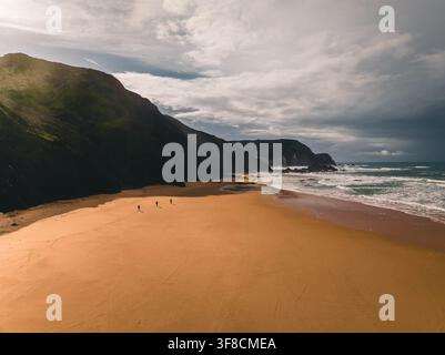 Vue aérienne de Praia da Cordoama avec sa plage de sable étendue, ses falaises dramatiques et ses vagues de l'océan, montrant trois personnes marchant le long du rivage à L Banque D'Images