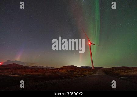 Vue d'une éolienne illuminée par des lumières d'avertissement rouges sous un spectacle vibrant d'aurore boréale dans le ciel nocturne au-dessus du paysage accidenté de Kvaloya Banque D'Images