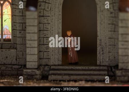 Une petite figurine de moine aux mains jointes se tient dans l'entrée sombre d'un modèle d'église en pierre à côté d'une vitre en vitrail lumineuse. Banque D'Images