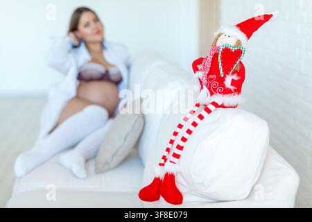 Un elfe en peluche doux est assis sur un canapé blanc avec une femme enceinte se détendant en arrière-plan flou. Banque D'Images