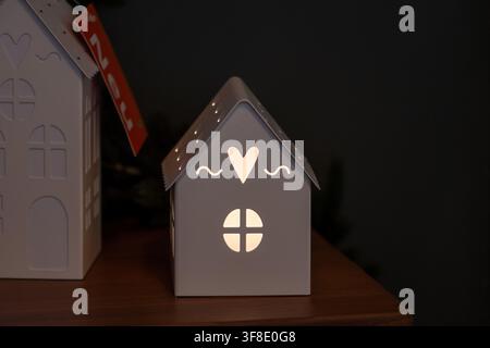 Une petite lanterne en forme de maison blanche avec des découpes en forme de cœur et de fenêtre brille sur une surface en bois devant un fond sombre. Banque D'Images