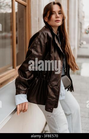 Veste en cuir pour femme style urbain brune portant une mode automnale tendance tout en s'appuyant sur le rebord d'une fenêtre de la ville Banque D'Images